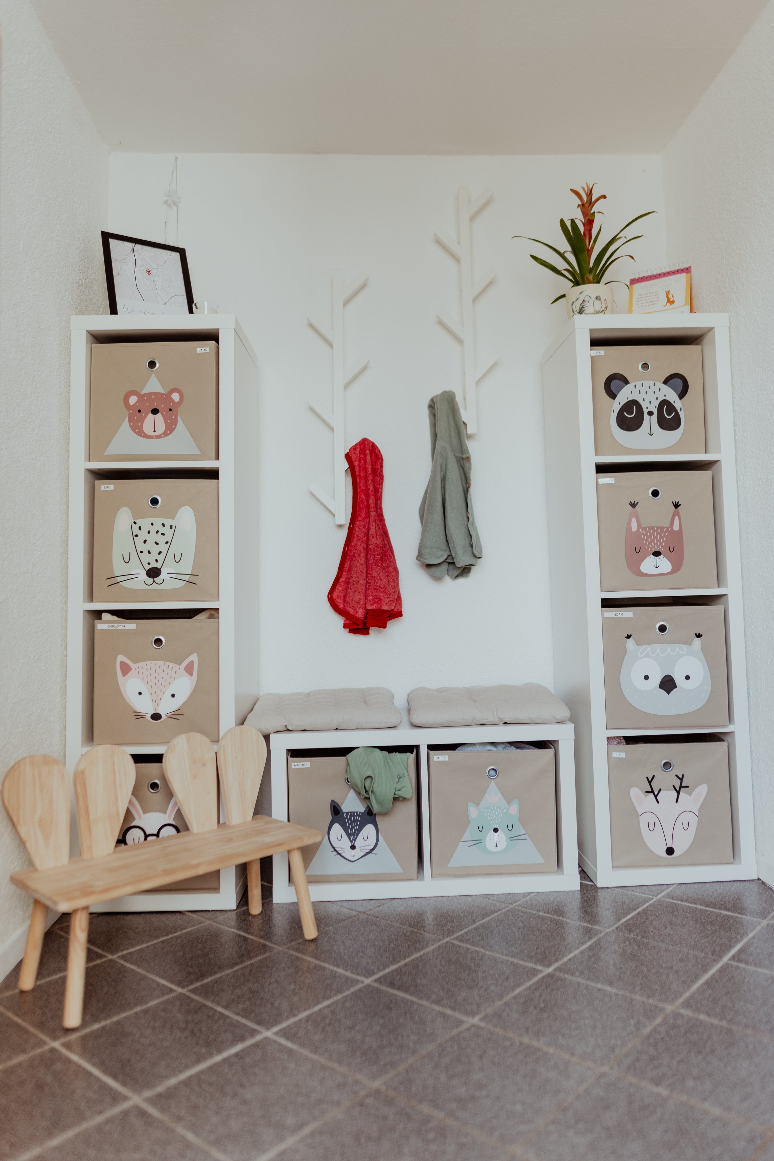 Kinderzimmer mit weißen Regalen, die mit Aufbewahrungskästen mit Tiergesichtern gefüllt sind, und einer kleinen Holzbank. Es hängen Handtücher an einer Wand mit Tiergesicht-Designs, darunter eine Pflanze und ein Bilderrahmen.