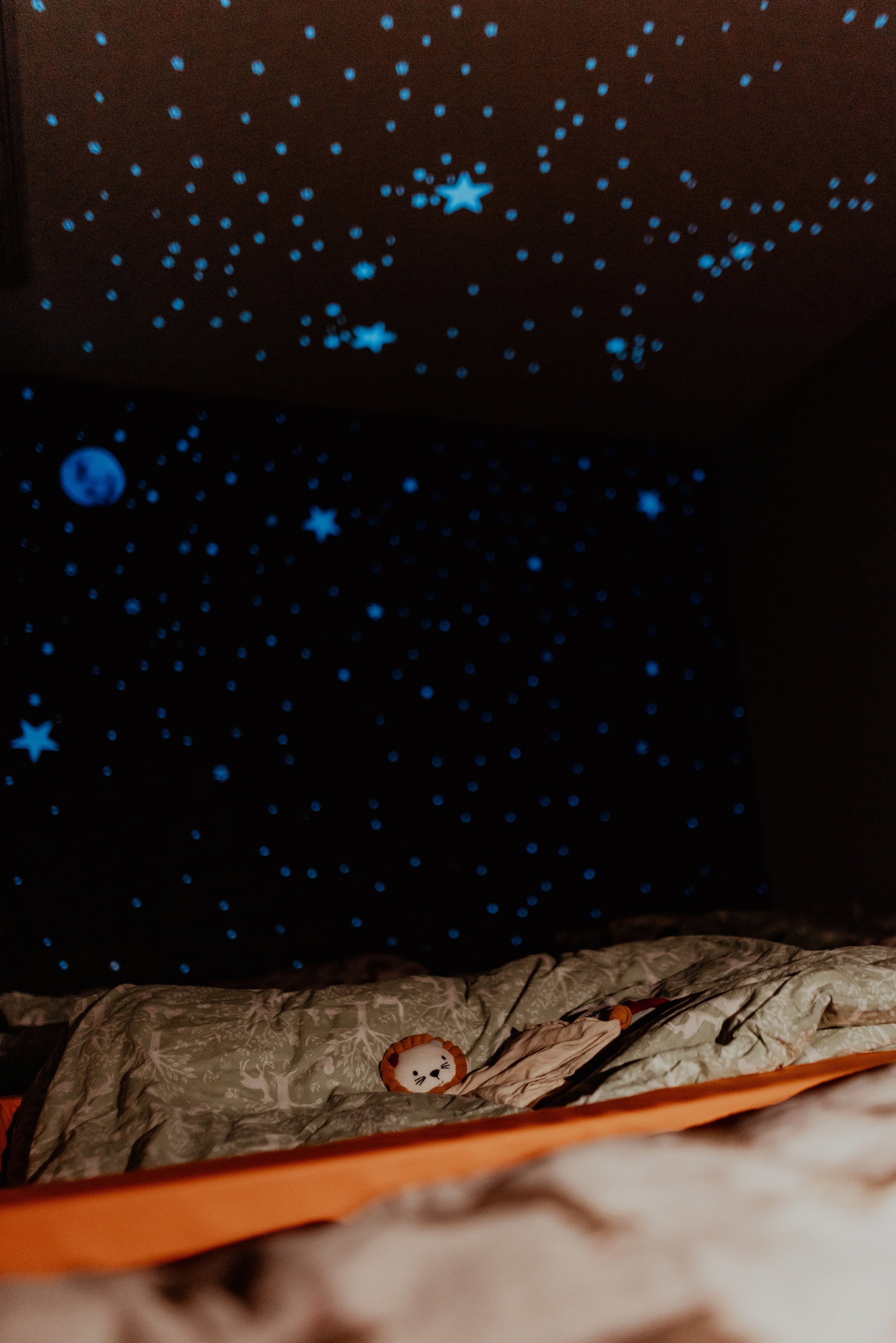 Schlafzimmer mit leuchtenden Sternen und Planeten an der Decke und Wand, Bett mit Decke und Kissen, kleine Plüschtiere auf dem Bett.