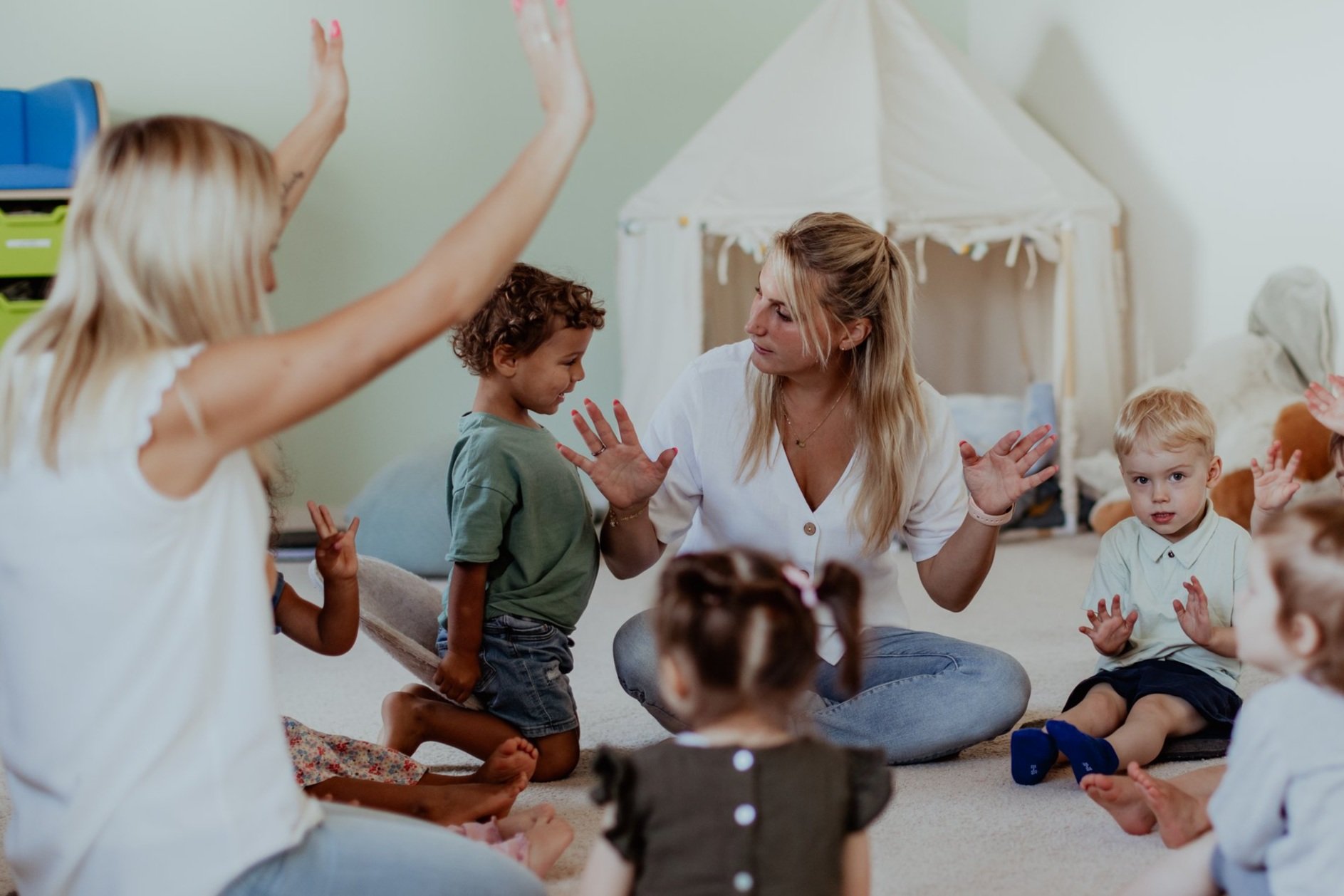 Eine Erzieherin sitzt im Kreis mit kleinen Kindern in einem Spielzimmer, einige Kinder haben die Hand gehoben, um zu antworten oder Fragen zu stellen.