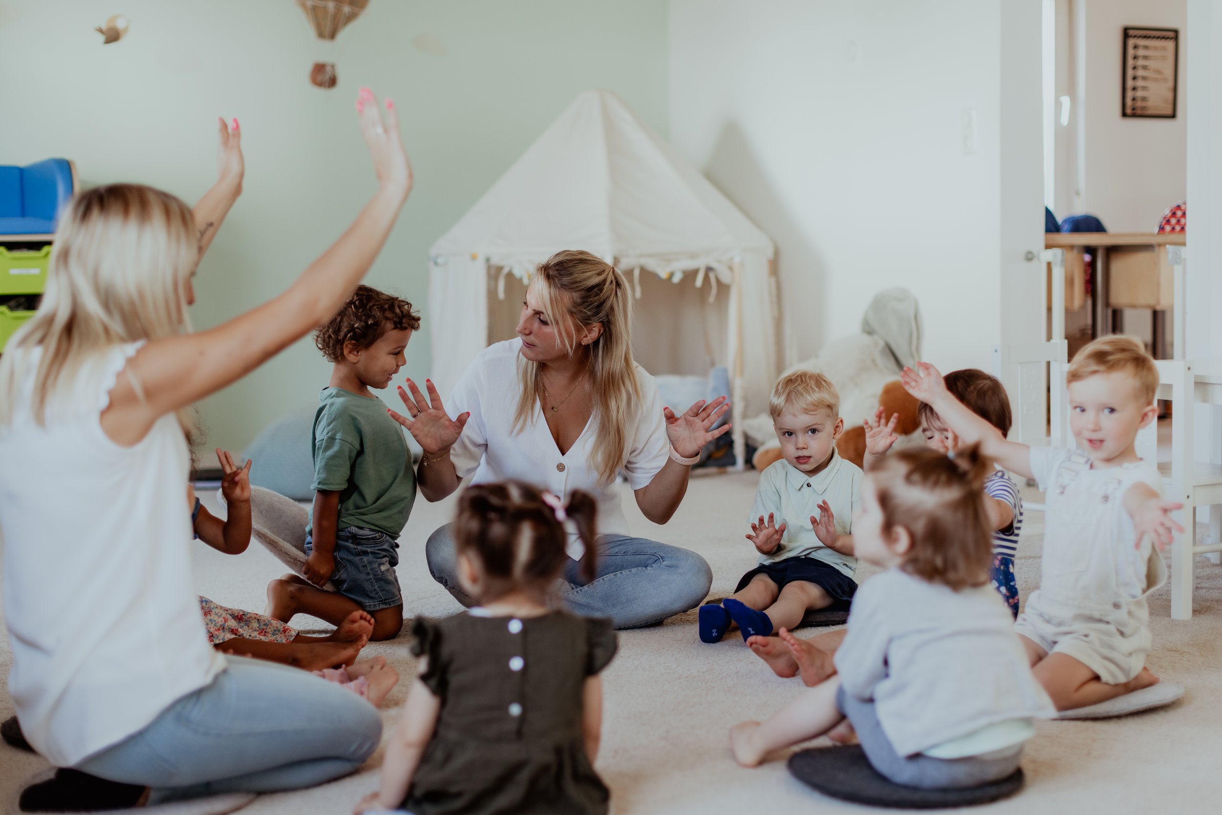 Eine Gruppe von Kindern sitzt im Kreis auf dem Boden, während eine Frau in der Mitte mit den Armen nach oben gestikuliert, vermutlich beim Singen oder Spielen in einem Kindertagesstättenraum.