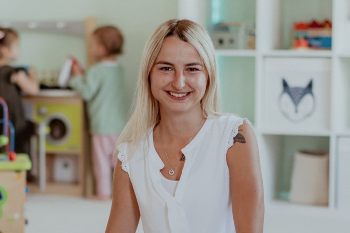Ein blonde Frau lächelt in einem Raum mit Spielzeugen, im Hintergrund sind Kinder und ein Regal mit bunten Gegenständen.