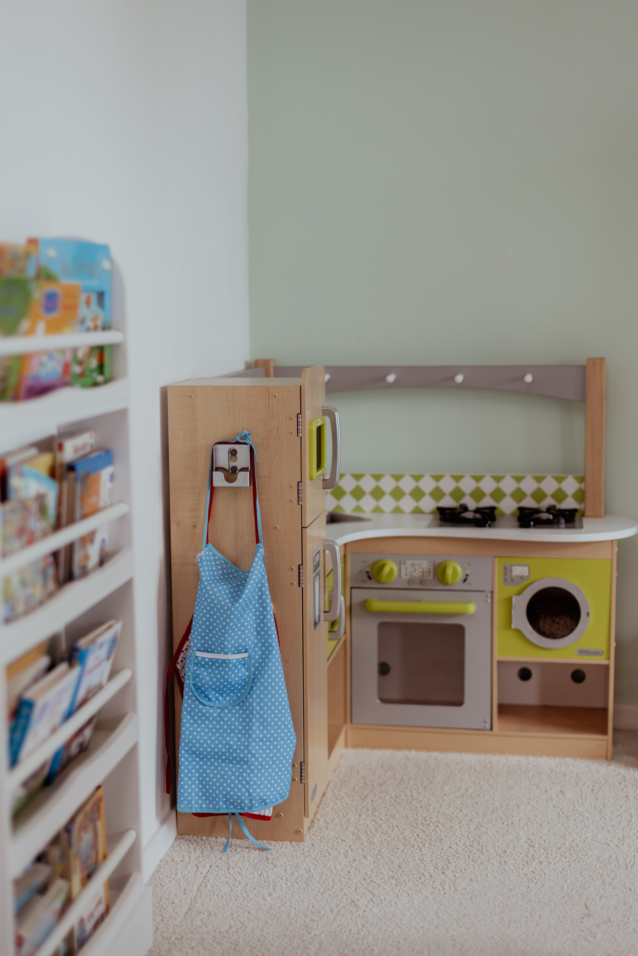 Kinderküche mit Spielzeugherd, Waschmaschine und kleinen Küchenutensilien, daneben ein Regal mit Kinderbüchern, im Vordergrund hängt eine blaue Tasche an der Küchenwand.