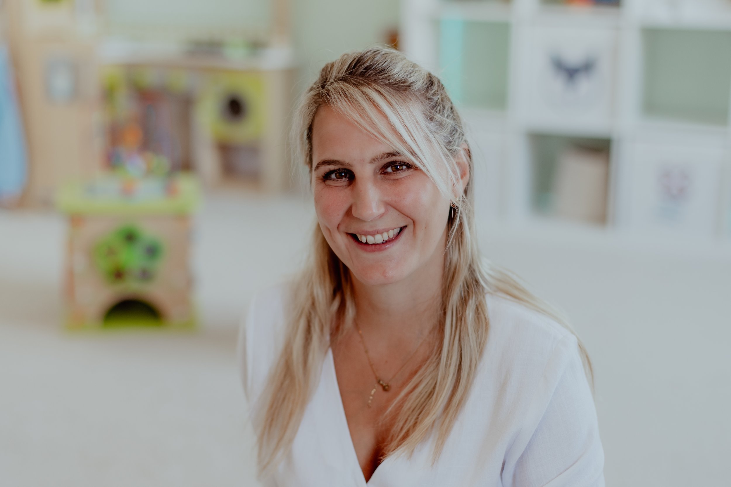 Lächelnde Frau in einem hellen Raum, vermutlich Kinderbetreuung oder Kindergarten, im Hintergrund Spielzeug und Regale.
